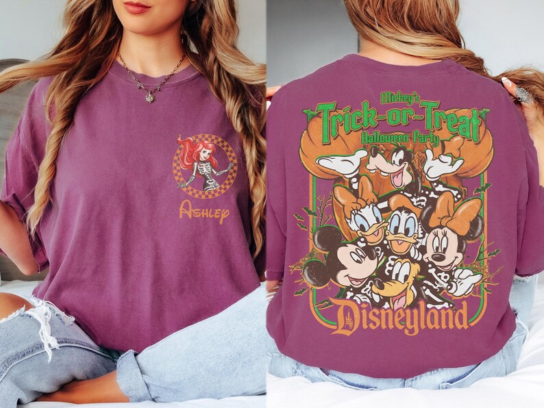 May include: A plum-colored t-shirt featuring a small circular graphic on the front with a skeleton-costumed character and the name Ashley. The back displays a large graphic of Disney characters in skeleton costumes with the text Mickey's Trick-or-Treat Halloween Party Disneyland.