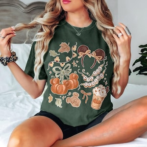 May include: A dark green t-shirt featuring a graphic print with orange pumpkins, brown mouse ears, a string of beads with the text 'TIS THE SEASON', and a cup of coffee with whipped cream, all accented with small leaves and decorative bows.