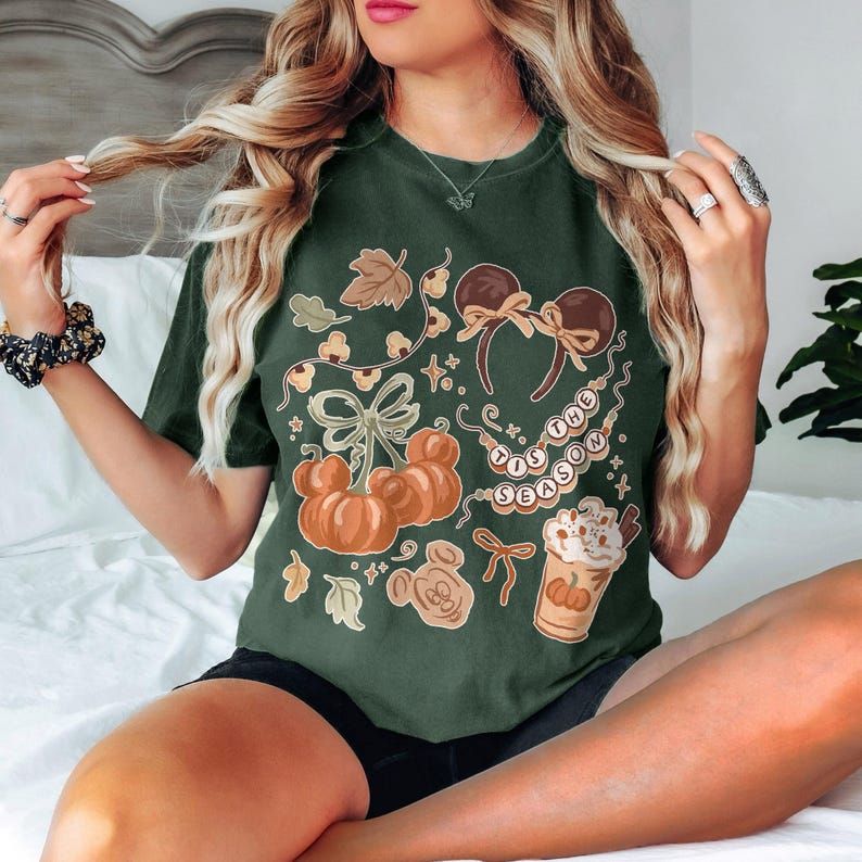May include: A dark green t-shirt featuring a graphic print with orange pumpkins, brown mouse ears, a string of beads with the text 'TIS THE SEASON', and a cup of coffee with whipped cream, all accented with small leaves and decorative bows.