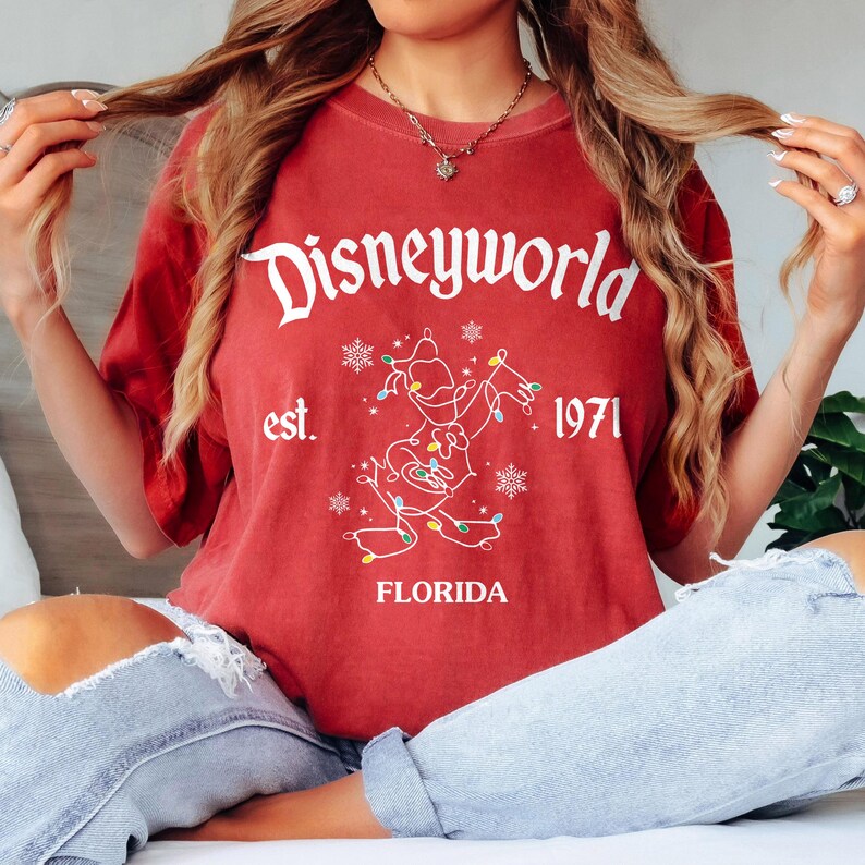 May include: A red t-shirt featuring the white text "Disneyworld" and a graphic of Mickey Mouse adorned with Christmas lights. The shirt also includes the text "est. 1971" and "Florida".