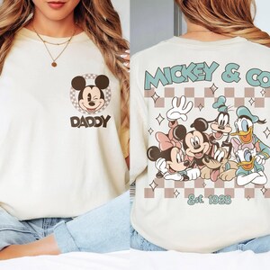 K&ouml;nnte beinhalten: Cremefarbenes T-Shirt mit Mickey Mouse und dem Wort "DADDY" darunter. Auf der R&uuml;ckseite des Shirts steht "Mickey & Co" mit verschiedenen Disney-Figuren und dem Text "Est. 1928" in einem Schachbrettmuster.