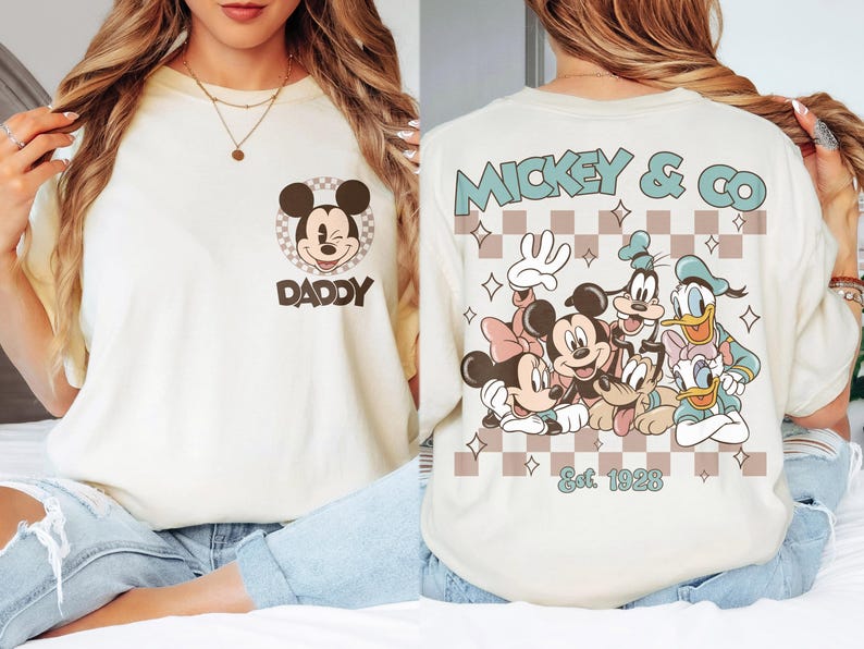 K&ouml;nnte beinhalten: Cremefarbenes T-Shirt mit Mickey Mouse und dem Wort "DADDY" darunter. Auf der R&uuml;ckseite des Shirts steht "Mickey & Co" mit verschiedenen Disney-Figuren und dem Text "Est. 1928" in einem Schachbrettmuster.