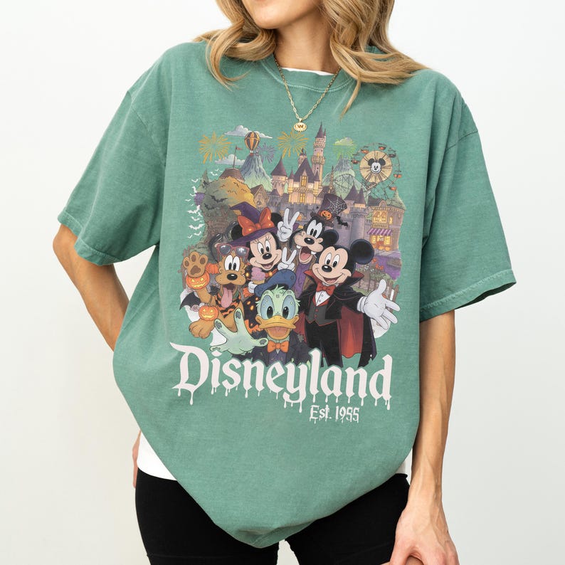 May include: A sage green short-sleeve t-shirt featuring a graphic print of Disney characters in Halloween costumes, including Mickey Mouse, Minnie Mouse, Donald Duck, and Pluto. The text reads Disneyland Est. 1955 with a dripping font effect.