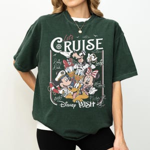 May include: Dark green t-shirt featuring a graphic of Disney characters, including Mickey Mouse, Minnie Mouse, Donald Duck, and Pluto. The shirt has the text "Let's Cruise" and "Disney Wish" in a white font.