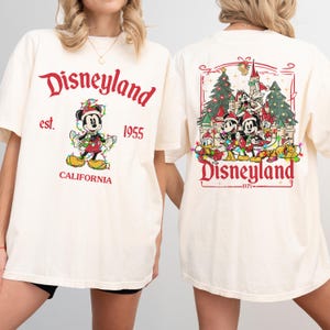May include: Cream-colored t-shirt with "Disneyland" in red script, featuring Mickey Mouse in a festive outfit. The back displays a Disneyland scene with characters and text. "Est. 1955 California" is printed on the front.