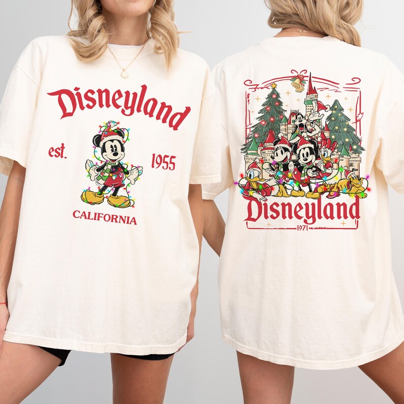 May include: Cream-colored t-shirt with "Disneyland" in red script, featuring Mickey Mouse in a festive outfit. The back displays a Disneyland scene with characters and text. "Est. 1955 California" is printed on the front.