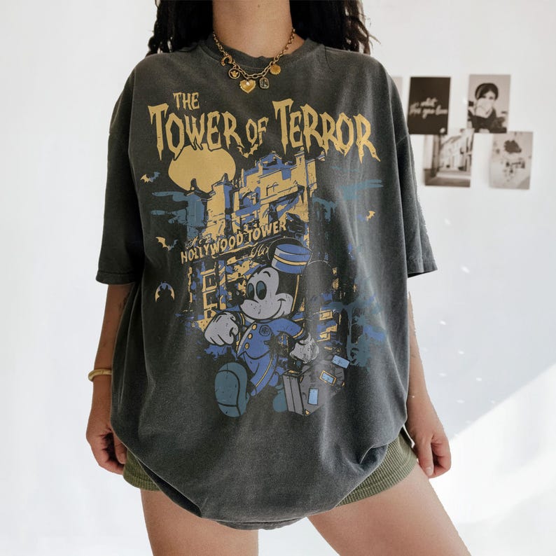 May include: Dark gray t-shirt featuring a graphic of Mickey Mouse in a bellhop uniform in front of the Hollywood Tower Hotel. The text "The Tower of Terror" is printed above the image in yellow and black.