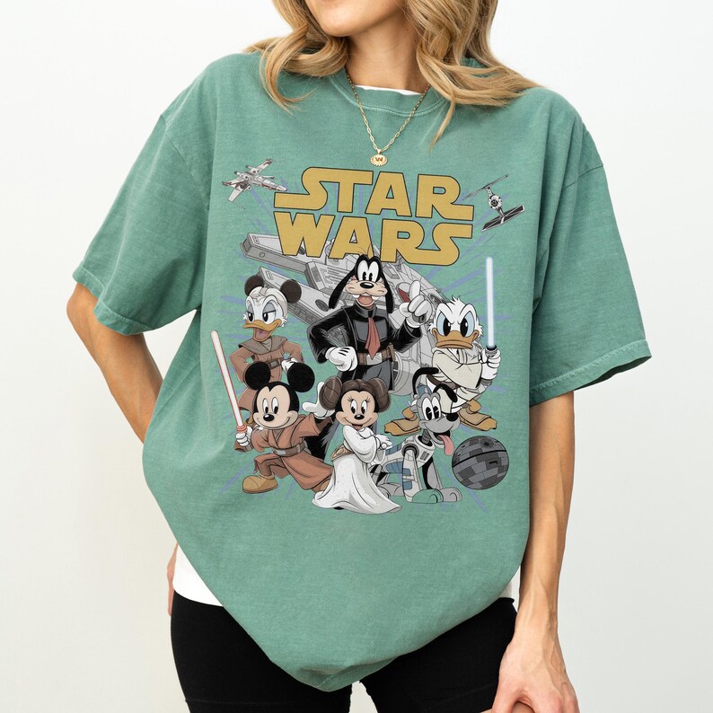 May include: Green t-shirt featuring a Star Wars graphic with Disney characters. The design includes Mickey Mouse, Minnie Mouse, Donald Duck, Goofy, and others, with lightsabers and a spaceship. The words "STAR WARS" are in gold.