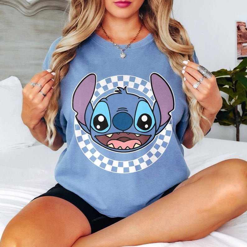 May include: A pale blue t-shirt with a cartoon image of Stitch from Lilo & Stitch. The image of Stitch is centered on the shirt, within a white and grey chequered circle. The shirt has short sleeves.
