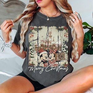 May include: Dark gray t-shirt featuring Mickey and Minnie Mouse in Christmas attire, holding a camera in front of a castle. The shirt has the words "Merry Christmas" in a script font. The design includes Christmas trees and candy canes.