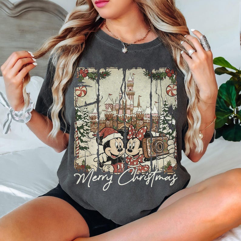 May include: Dark gray t-shirt featuring Mickey and Minnie Mouse in Christmas attire, holding a camera in front of a castle. The shirt has the words "Merry Christmas" in a script font. The design includes Christmas trees and candy canes.