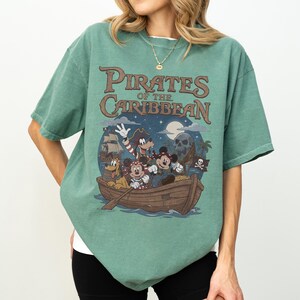 K&ouml;nnte beinhalten: Gr&uuml;nes T-Shirt mit einem Pirates of the Caribbean-Design. Die Grafik zeigt Zeichentrickfiguren in einem Boot, ein Piratenschiff, einen Totenkopf und den Text "Pirates of the Caribbean".