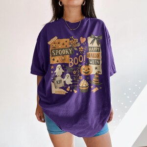 K&ouml;nnte beinhalten: Ein lila T-Shirt mit einem Halloween-Motiv. Das Design enth&auml;lt die W&ouml;rter "Spooky Boo! Happy Halloween" zusammen mit Bildern von Geistern, K&uuml;rbissen, einem Hexenhut und einem Cupcake.