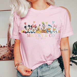 Puede incluir: Camiseta rosa claro con un gr&aacute;fico colorido de Mickey Mouse, Minnie Mouse, Pluto, Goofy, Donald Duck y Daisy Duck. El texto "MICKEY & CO Est. 1928" est&aacute; impreso debajo. La camiseta tiene mangas cortas.