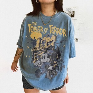 May include: Blue t-shirt featuring a graphic of Mickey Mouse in a bellhop uniform, the Hollywood Tower Hotel, and the text "The Tower of Terror." The design is in shades of yellow, blue, and gray.