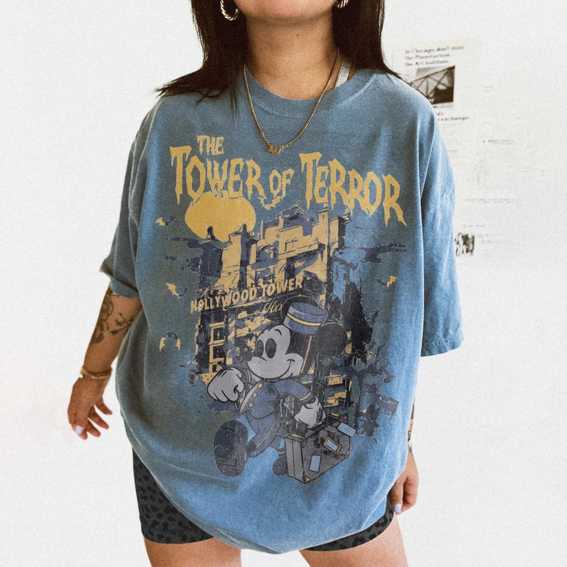 May include: Blue t-shirt featuring a graphic of Mickey Mouse in a bellhop uniform, the Hollywood Tower Hotel, and the text "The Tower of Terror." The design is in shades of yellow, blue, and gray.