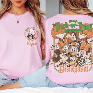 May include: A light pink t-shirt featuring a small graphic of Marie from The Aristocats on the front left chest with the name Kayla below. The back displays a large graphic of Mickey Mouse and friends in skeleton costumes with the text Mickey's Trick-or-Treat Halloween Party Disneyland.