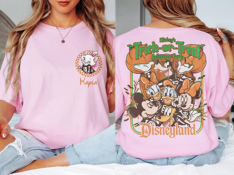 May include: A light pink t-shirt featuring a small graphic of Marie from The Aristocats on the front left chest with the name Kayla below. The back displays a large graphic of Mickey Mouse and friends in skeleton costumes with the text Mickey's Trick-or-Treat Halloween Party Disneyland.