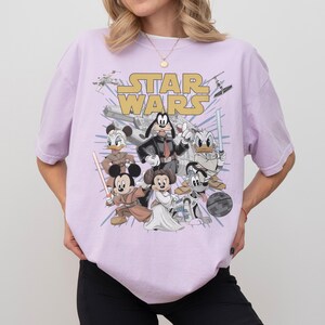 May include: A light purple t-shirt featuring a graphic of Disney characters dressed as Star Wars characters. The words "STAR WARS" are printed in gold across the top. Characters include Mickey Mouse, Donald Duck, and Goofy.