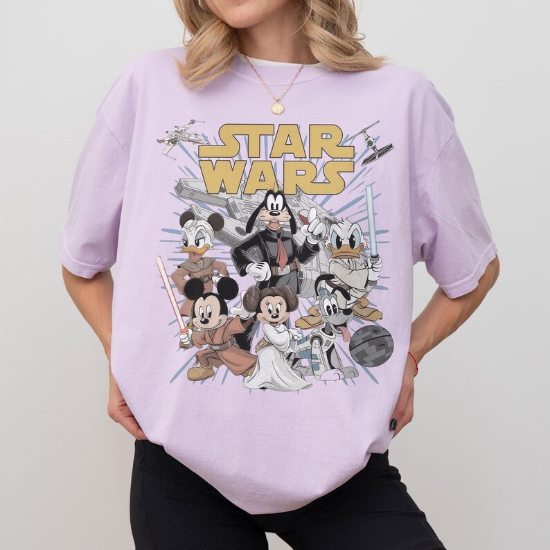 May include: A light purple t-shirt featuring a graphic of Disney characters dressed as Star Wars characters. The words "STAR WARS" are printed in gold across the top. Characters include Mickey Mouse, Donald Duck, and Goofy.