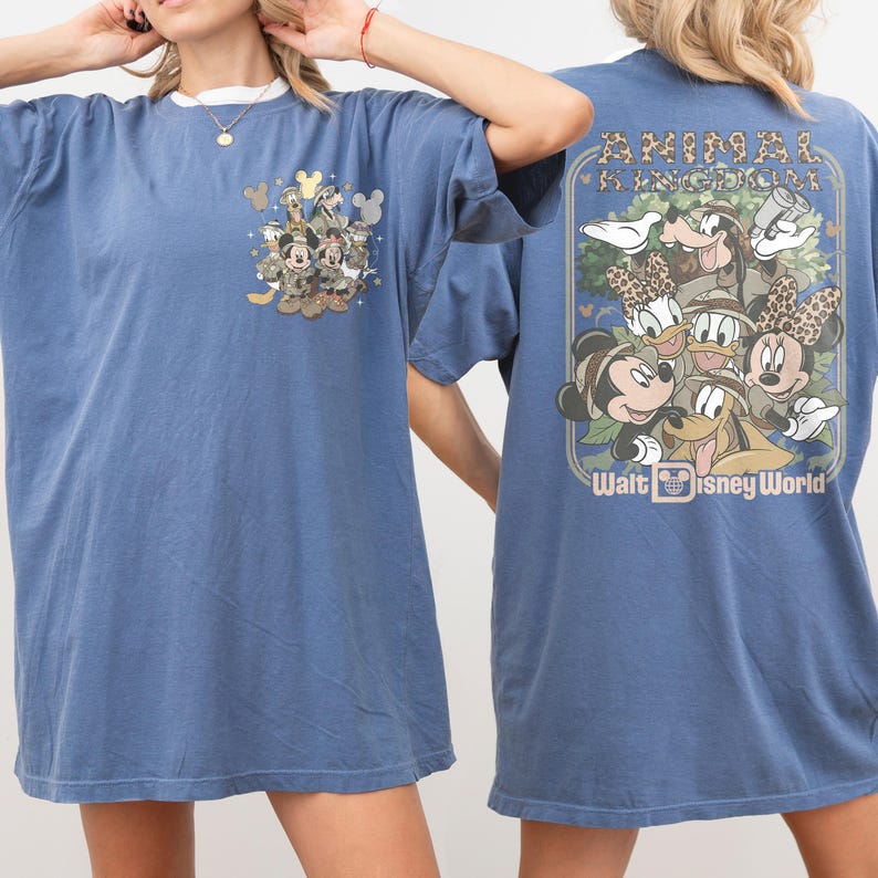 Puede incluir: Camiseta azul con un estampado de personajes de Disney, incluyendo Mickey Mouse, Minnie Mouse, Donald Duck y Goofy. La camiseta tiene el texto "Animal Kingdom" y "Walt Disney World". La camiseta tiene un cuello blanco.