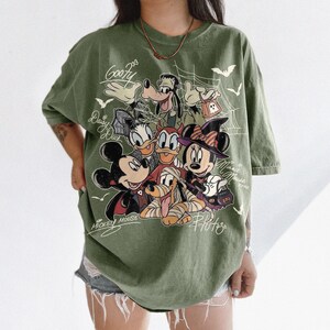 Może przedstawiać: Oliwkowa koszulka z nadrukiem graficznym przedstawiającym postacie Disneya w kostiumach na Halloween, w tym Myszkę Miki, Myszkę Minnie, Goofy'ego, Kaczora Donalda, Daisy i Pluto, z pajęczynami, nietoperzami i odręcznie napisanymi imionami.