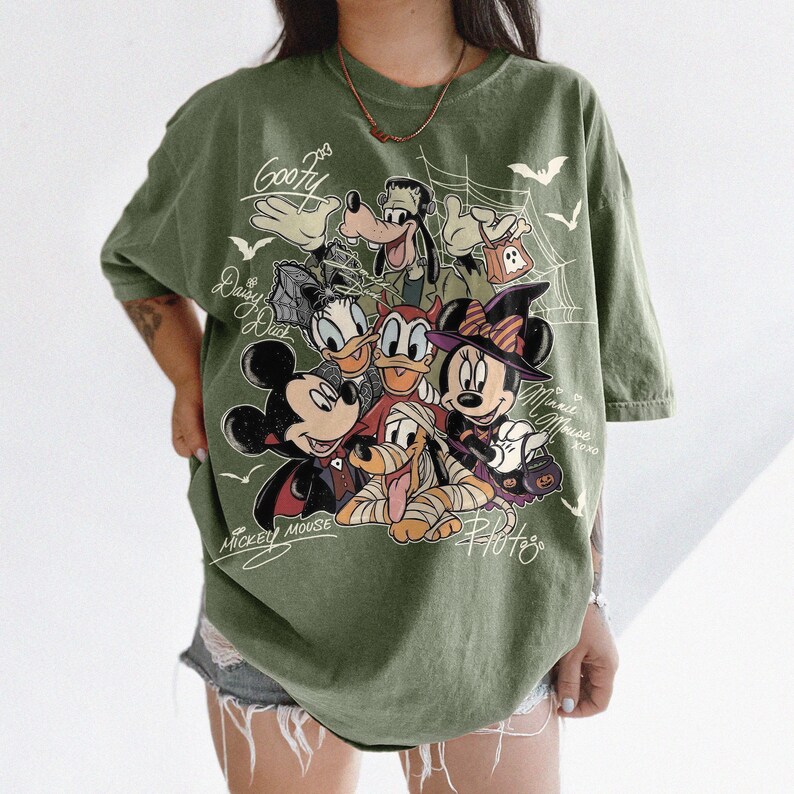 Może przedstawiać: Oliwkowa koszulka z nadrukiem graficznym przedstawiającym postacie Disneya w kostiumach na Halloween, w tym Myszkę Miki, Myszkę Minnie, Goofy'ego, Kaczora Donalda, Daisy i Pluto, z pajęczynami, nietoperzami i odręcznie napisanymi imionami.