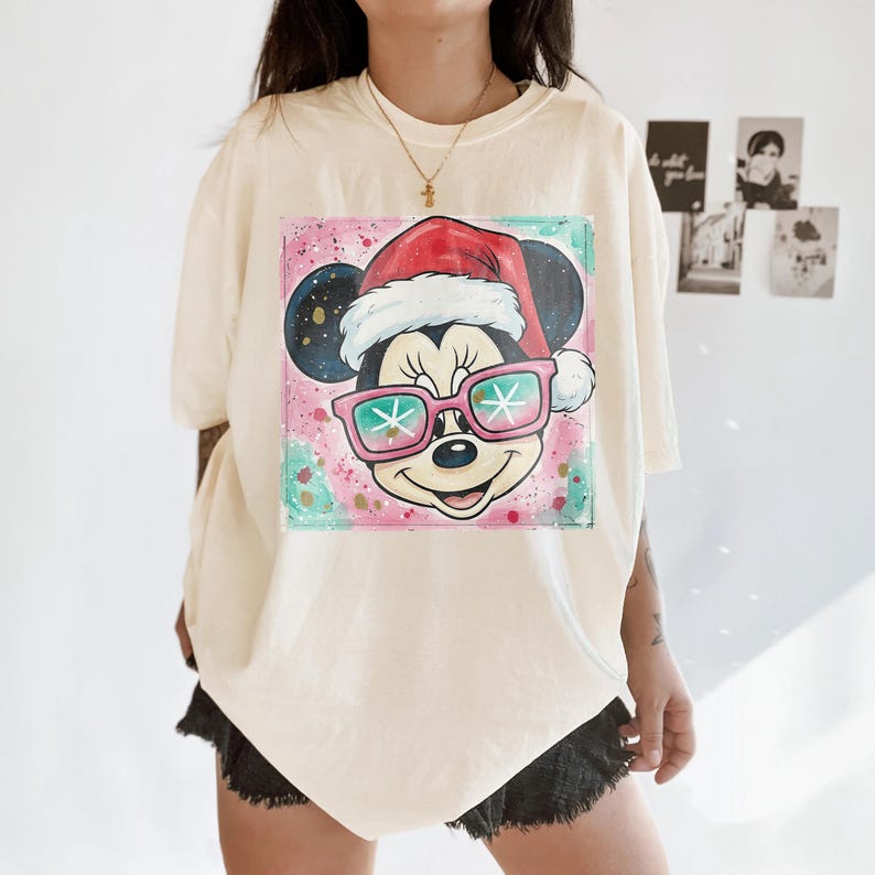 May include: Cream-colored t-shirt featuring a colorful graphic of Mickey Mouse wearing a Santa hat and pink sunglasses with snowflake designs. The background has pink, blue, and green paint splatters. A gold necklace is visible.