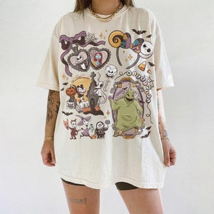 May include: Cream-coloured t-shirt featuring a colourful graphic design with characters and elements from "The Nightmare Before Christmas." The design includes Jack Skellington, Sally, Oogie Boogie, and other Halloween-themed imagery. The shirt has short sleeves and a relaxed fit.
