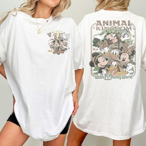 Puede incluir: Camiseta blanca con dise&ntilde;os gr&aacute;ficos en la parte delantera y trasera. El dise&ntilde;o frontal incluye personajes de dibujos animados en camuflaje. El dise&ntilde;o trasero dice "ANIMAL KINGDOM" y "Walt Disney World" con personajes de dibujos animados con atuendo de safari.