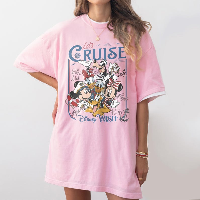 May include: A light pink t-shirt featuring a graphic of Disney characters, including Mickey Mouse, Minnie Mouse, and Donald Duck. The shirt has the words "Let's Cruise" and "Disney Wish" printed on it. The shirt has short sleeves and a white trim.