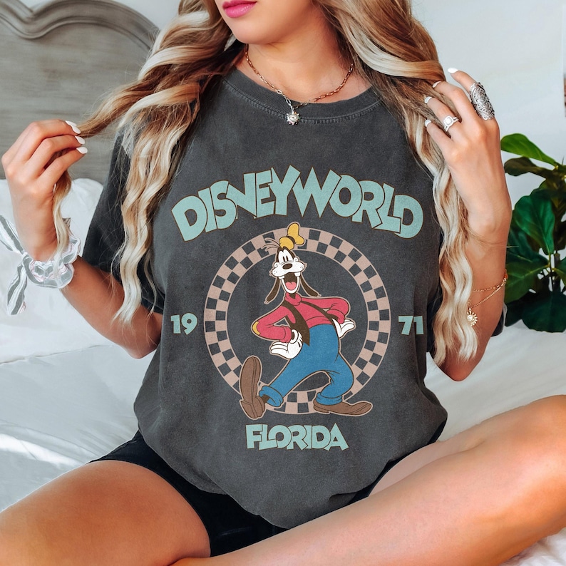 May include: Dark gray t-shirt featuring a vintage Disney World design. The design includes the text "DISNEYWORLD", "1971", and "FLORIDA", along with a cartoon image of Goofy. The shirt has a classic, worn look.