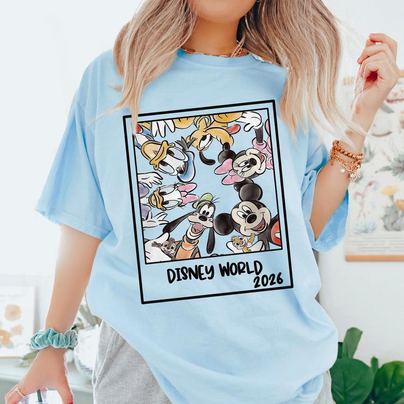 Puede incluir: Camiseta azul claro con un gr&aacute;fico enmarcado de personajes de Disney, incluyendo Mickey Mouse, Minnie Mouse, Donald Duck y Goofy. El gr&aacute;fico est&aacute; rodeado por un borde negro y tiene el texto "DISNEY WORLD 2026".