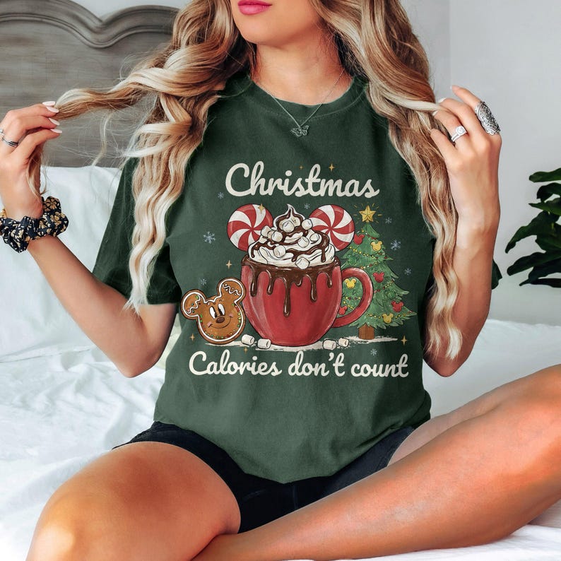 May include: Dark green t-shirt with a Christmas-themed graphic. The design features a red mug of hot chocolate with whipped cream, candy cane ears, and a gingerbread cookie. The text "Christmas Calories don't count" is also included.