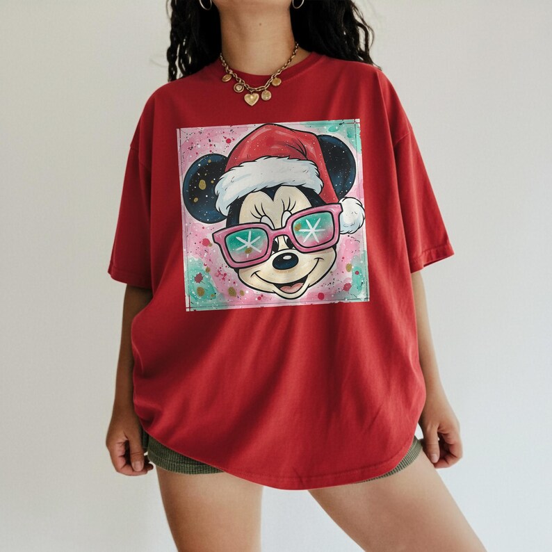 May include: Red t-shirt featuring a graphic of Mickey Mouse wearing a Santa hat and pink sunglasses. The graphic is set against a pink, green, and white background. The shirt has short sleeves and a relaxed fit.