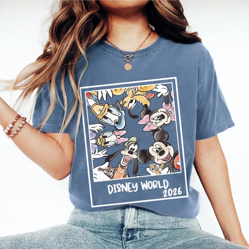 Puede incluir: Camiseta azul con un dise&ntilde;o de personajes de Disney, incluyendo Mickey y Minnie Mouse, Donald Duck y Goofy. El dise&ntilde;o est&aacute; enmarcado en blanco y tiene el texto "Disney World 2026".