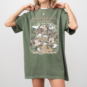 May include: Olive green t-shirt featuring a graphic of Disney characters, including Mickey and Minnie Mouse, Donald and Daisy Duck, Goofy, and Pluto. The shirt has the text "ANIMAL KINGDOM" and "Walt Disney World" in a vintage style.