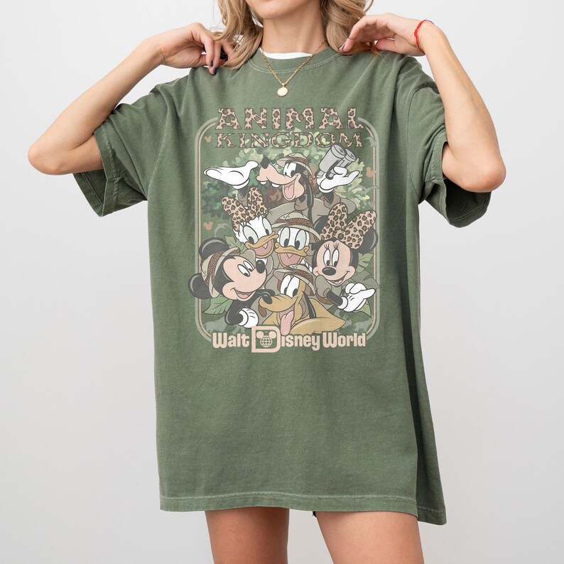 May include: Olive green t-shirt featuring a graphic of Disney characters, including Mickey and Minnie Mouse, Donald and Daisy Duck, Goofy, and Pluto. The shirt has the text "ANIMAL KINGDOM" and "Walt Disney World" in a vintage style.