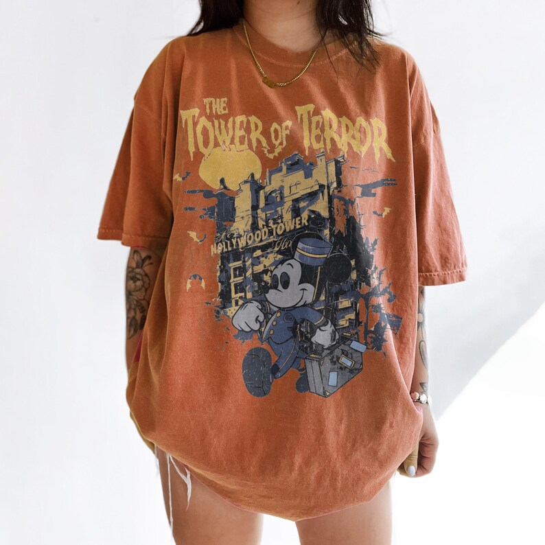 May include: Orange t-shirt featuring a graphic of Mickey Mouse in a bellhop uniform, with the text "The Tower of Terror" and "Hollywood Tower". The design is distressed, giving the shirt a vintage look.
