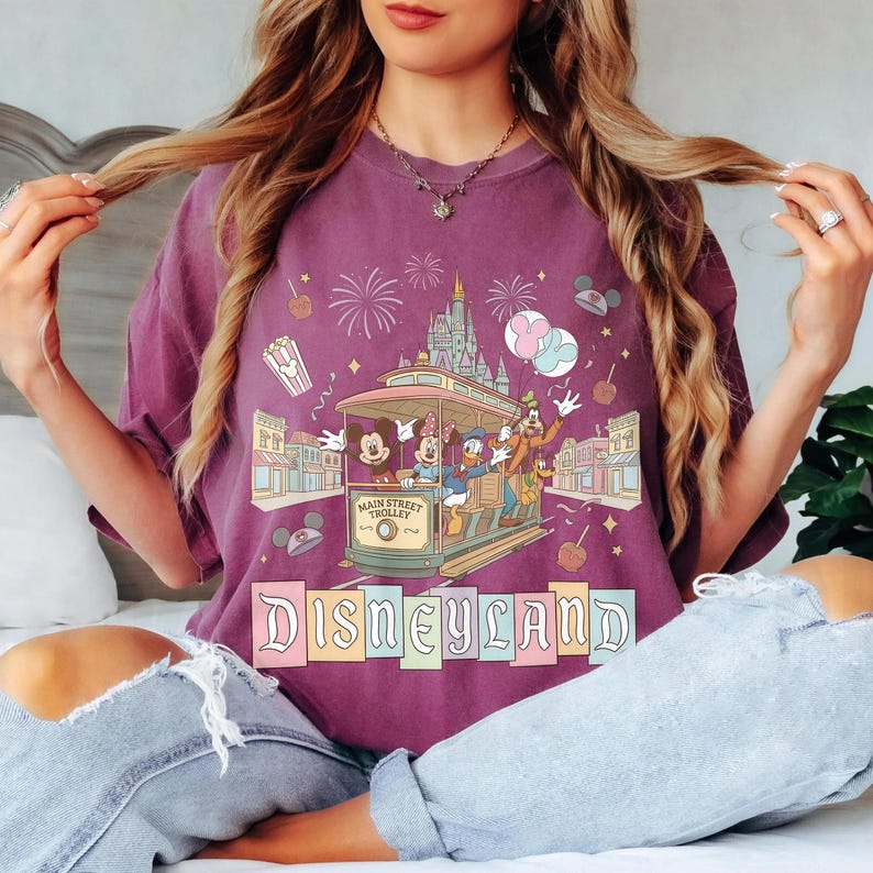 K&ouml;nnte beinhalten: Ein lila T-Shirt mit einem Disneyland Main Street Trolley-Design mit Micky Maus, Minnie Maus, Donald Duck und Goofy. Das Design umfasst Feuerwerk, ein Schloss und das Wort "Disneyland".