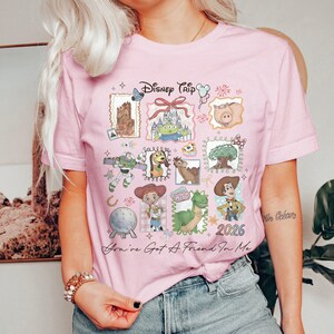 May include: Pink t-shirt with a colorful graphic featuring Disney characters and the text "Disney Trip 2026". The design includes images of Buzz Lightyear, Woody, and other characters from the Toy Story franchise.