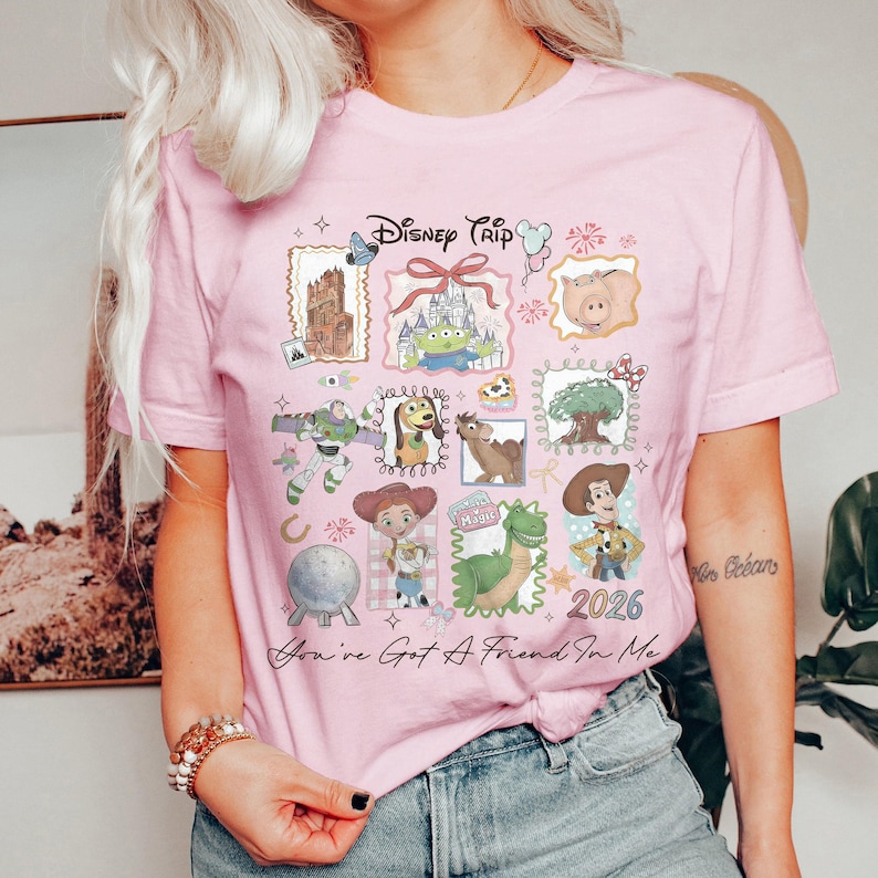 May include: Pink t-shirt with a colorful graphic featuring Disney characters and the text "Disney Trip 2026". The design includes images of Buzz Lightyear, Woody, and other characters from the Toy Story franchise.