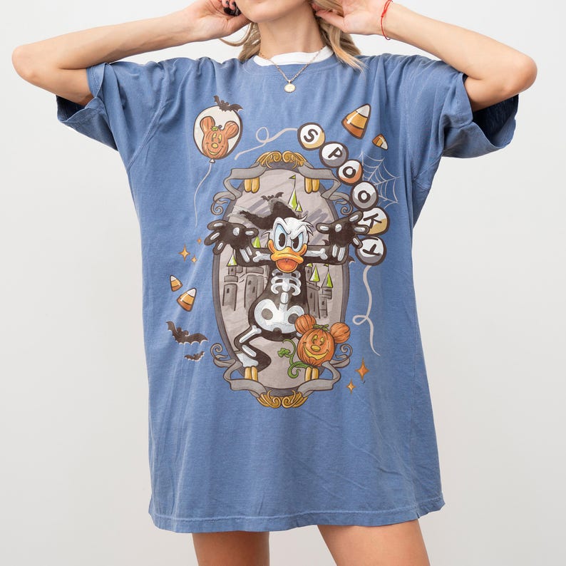 May include: Blue t-shirt featuring a Halloween-themed graphic of Donald Duck as a skeleton. The design includes a castle, bats, candy corn, and the word "SPOOKY". The shirt has short sleeves and a crew neck.