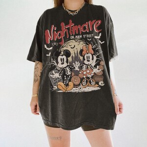 May include: Dark gray t-shirt with a Halloween-themed graphic. The design features Mickey and Minnie Mouse as skeletons in front of a spooky castle, with the words "Nightmare on Main Street" in red and white.