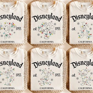 May include: Six folded cream-colored t-shirts with the Disneyland logo, Mickey Mouse, Pluto, and Goofy characters, and the text "est. 1955 CALIFORNIA". The shirts are arranged on woven mats.
