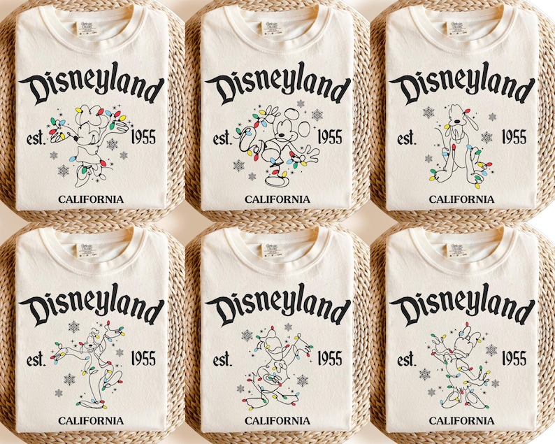 May include: Six folded cream-colored t-shirts with the Disneyland logo, Mickey Mouse, Pluto, and Goofy characters, and the text "est. 1955 CALIFORNIA". The shirts are arranged on woven mats.