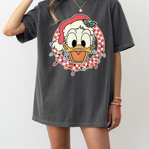 May include: Dark gray t-shirt featuring a cartoon Donald Duck wearing a Santa hat and a wreath of Christmas lights. The wreath has a red and white checkered pattern. The shirt is a relaxed fit.