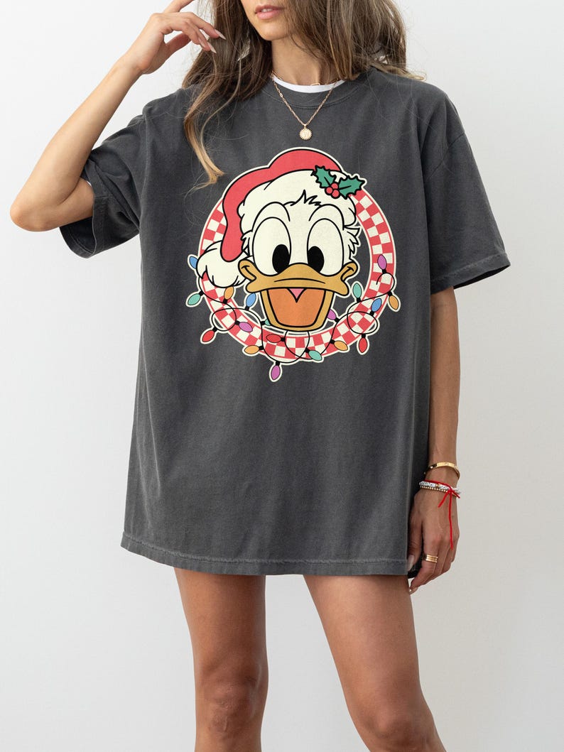 May include: Dark gray t-shirt featuring a cartoon Donald Duck wearing a Santa hat and a wreath of Christmas lights. The wreath has a red and white checkered pattern. The shirt is a relaxed fit.