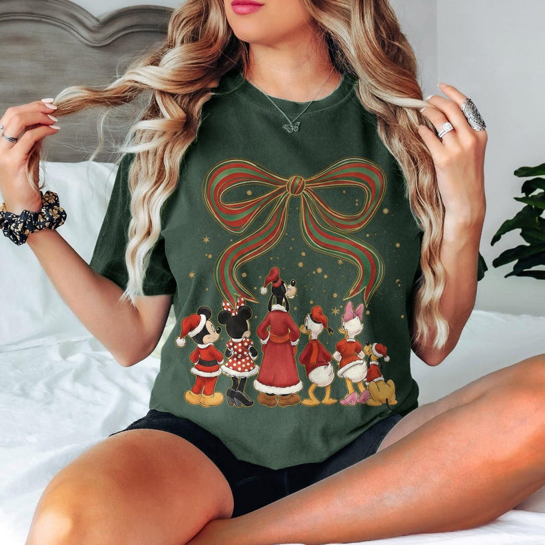 May include: Dark green t-shirt featuring a large red and gold bow design with a group of cartoon characters in Christmas attire. The characters include Mickey and Minnie Mouse, Donald and Daisy Duck, and Goofy. The shirt is a comfortable, casual style.