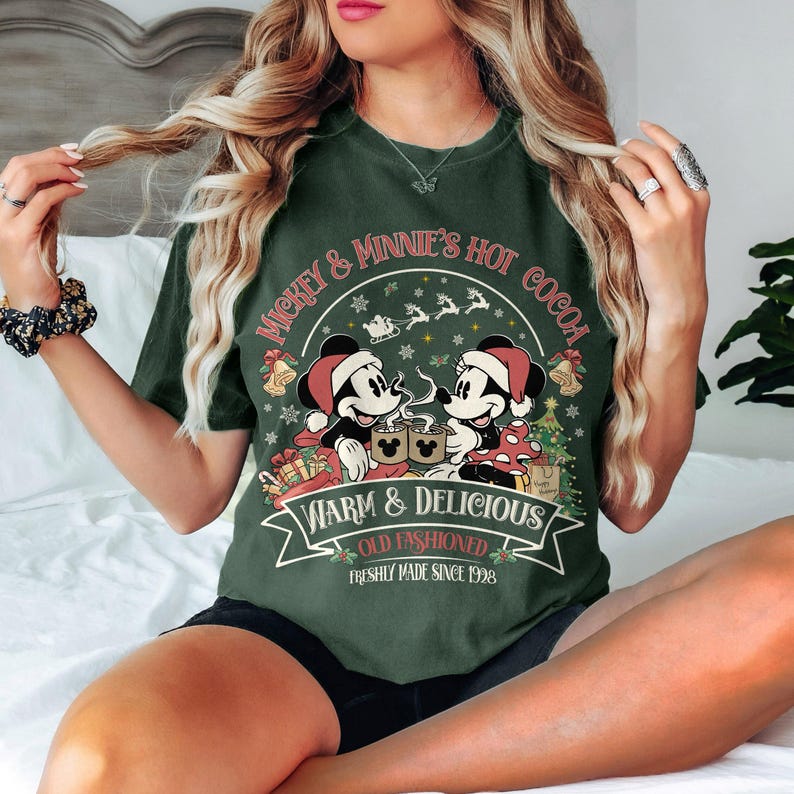May include: Dark green t-shirt featuring Mickey and Minnie Mouse enjoying hot cocoa. The design includes the text "Mickey & Minnie's Hot Cocoa" and "Warm & Delicious". Festive elements like a Christmas tree and Santa hats are also present.
